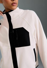 Fleecejacke mit hohem Kragen, Knopföffnung und kontrastierenden schwarzen Akzenten, einschließlich einer Reißverschlusstasche und Seitentaschen. Die Farbe ist hellrosa.