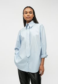 Ανοιχτό μπλε πουκάμισο με κλειστό γιακά, με κρεμασμένο φιόγκο, μακριά μανίκια διπλωμένα, λεία υφή και διακριτική επωνυμία στο κάτω μέρος.