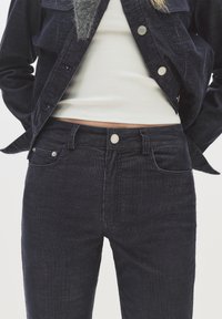 Ženska oblečena v temno modre kordne hlače in ujemajoči se jopič nad belim rebrastim kratkim topom, roke zadaj, proti enotni ozadju.