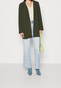 Blazer de corte oversized en verde oliva, combinado con una blusa beige de escote en V, jeans de pierna ancha en azul claro y botines en teal. Incluye un bolso verde.