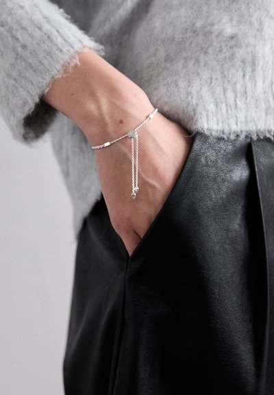 Bratara argintie cu un design de lanț împletit, având un charm circular și două capete suspendate, purtată pe o mână deasupra unei perechi de pantaloni din piele neagră.