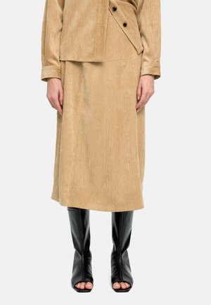 Kobieta ubrana w beżowy zestaw z sztruksowej spódnicy i kurtki, w połączeniu z czarnymi, otwartymi u góry, wysokimi na kolano butami, stojąca na neutralnym tle.