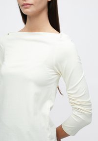 Maglia a maniche lunghe color crema con scollo ampio e spalle arricciate. Tessuto morbido con texture liscia, silhouette aderente.