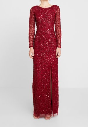 Abito lungo fino a terra con maniche lunghe, aderente, di colore rosso con decorazioni di paillettes e spacco frontale, indossato con tacchi aperti.