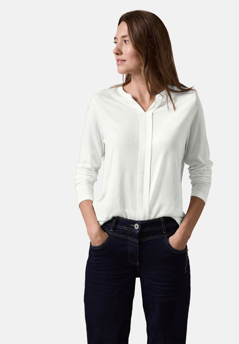 Valkoinen pitkähiha blouse, jossa on V-mallinen kaula-aukko ja etupaneeli, yhdistetty tummansinisiin, hieman teksturoituihin farkkuihin, joissa on näkyvät tikit ja taskut.