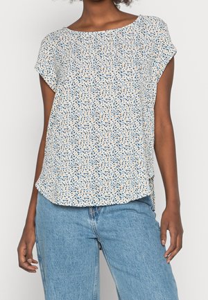 Vrouw draagt een wit blouse met korte mouwen met kleine blauwe en perzikkleurige stippen, gecombineerd met lichtblauwe denim jeans.