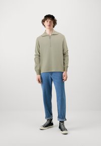 Lys grønn genser med halv-zip krage, paret med avslappede blå jeans og svarte høye sneakers. Glatt tekstur, uformell passform.