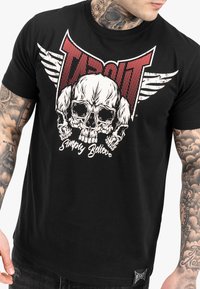 Schwarzes Baumwoll-T-Shirt mit einem zentralen Totenkopf-Graphic, roten und weißen Flügeldetails sowie dem Text "Simply Believe" und einem Logotag am Saum.