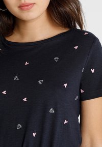 Černé tričko s krátkým rukávem, zdobené rozptýlenou výšivkou růžových srdcí a diamantů. Měkký bavlněný materiál s uvolněným výstřihem.