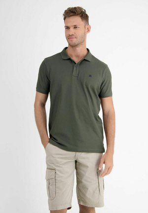 Mann trägt grünes Kurzarm-Poloshirt und beige Cargo-Shorts, steht mit einer Hand in der Tasche vor einem schlichten hellen Hintergrund.