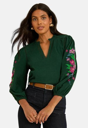 Blusa verde scuro con scollatura a V, impreziosita da ricami floreali colorati sulle maniche. Tessuto morbido, vestibilità comoda e maniche a sbuffo.