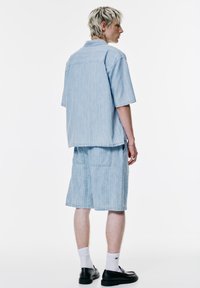 Camicia a maniche corte a righe blu chiaro e pantaloni corti coordinati, realizzati in tessuto morbido, con due tasche posteriori e vestibilità rilassata. Indossata con scarpe nere.
