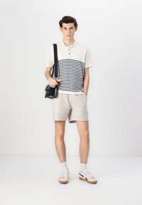 Polo à manches courtes avec des rayures navy, shorts beiges et baskets blanches. Sac à bandoulière noir. Fond clair.