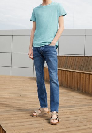 Light blue cotton t-shirt, blue jeans, and beige sandals with straps. Model stands on wooden deck against a gray backdrop.