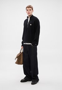 Čierny pletený sveter na zips s malým emblémom, nosený cez biele tričko. Kombinovaný so širokými tmavými rifľami a čiernymi topánkami. Drží hnedú tašku.