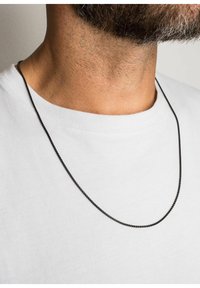 Sort metal kædelynke med et glat, delikat design, hvilende på en hvid T-shirt, synlig mod nakke- og brystområde.