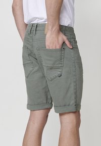 Shorts vert olive avec un ourlet roulé, plusieurs poches et un tissu texturé. Détails de couture notables et une patch en cuir sur la taille.
