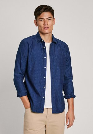 Camisa azul marino de botones con un sutil patrón de puntos; mangas largas arremangadas en los puños; llevada sobre una camiseta blanca. Material de algodón.