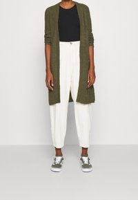 Oliivivihreä pitkä neuletakki, musta istuva paita, valkoiset kapeutetut housut ja oliivivihreät tennarit valkoisilla yksityiskohdilla. Neuletakissa on neulepinta.