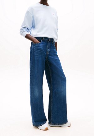 Persoon draagt lichtblauwe trui, wijde hoge taille spijkerbroek in blauw, witte sneakers, met één hand in de zak, staand voor een witte achtergrond.