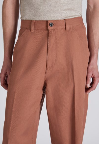 Pantaloni color ruggine con texture a costine diagonali, dotati di chiusura con bottone e tasche laterali. Vestibilità sartoriale con una finitura liscia.