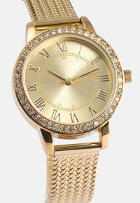 Χρυσό πλέγμα ρολογιού χειρός με στρογγυλό, βουρτσισμένο χρυσό καντράν. Διαθέτει ρομανικά αριθμητικά σημάδια και μπάντα διακοσμημένη με διαφανή κρύσταλλα γύρω από το περιθώριο.