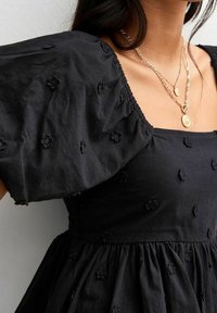 Robe en coton noire à manches courtes bouffantes, ornée de broderies florales et d'une taille froncée. Accessoirisée avec des colliers en or superposés.