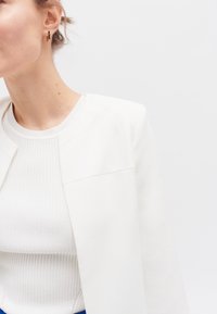 Witte gestructureerde blazer met een schone halslijn, gedragen over een witte geribbelde top, met minimale details en een gladde textuur.