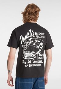 Camiseta de algodón negra con una impresión gráfica blanca de un tocadiscos y notas musicales, con texto sobre una tienda de discos en la parte de atrás.