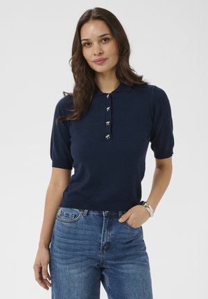Woman with long brown hair wearing a navy short-sleeve knit polo shirt with small bow buttons and blue jeans, hand in pocket.