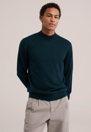 Jeune homme portant un pull vert foncé à manches longues et un pantalon beige clair, debout les mains dans le dos devant un fond uni.