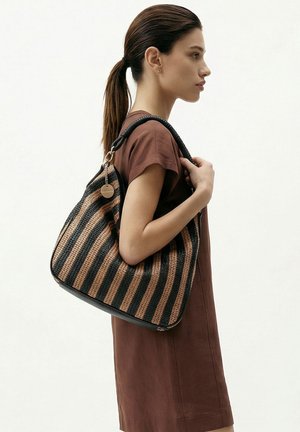 Femme en robe marron portant un grand sac tissé rayé noir et beige sur l'épaule, tournée vers la droite, les cheveux attachés en queue de cheval.