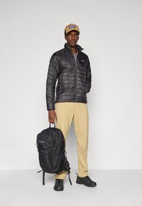 Veste noire isolée avec un motif matelassé, pantalon khaki, sac à dos noir avec logo, et chaussures foncées. Le modèle se tient devant un fond neutre.