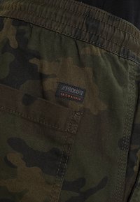 Kargo šortky z maskovacího materiálu v tmavě zelených tónech, s nášivkou s nápisem "//PRODUKT" a několika detaily kapes.