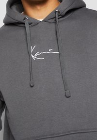 Personne portant un sweat à capuche gris avec des cordons et un logo en texte cursif blanc sur la poitrine.