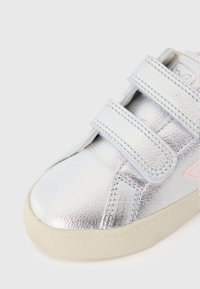 Μεταλλικό ασημί αθλητικό παπούτσι με δύο λουριά βέλκρο, επάνω μέρος από υφή δέρματος, ροζ λεπτομέρεια και λευκή ελαστική σόλα. Στρογγυλεμένη μύτη.