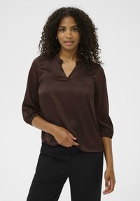 Blusa de satén marrón con escote en V y detalle de volantes, mangas 3/4 con puños elásticos, llevada sobre pantalones negros. Textura suave, corte holgado.