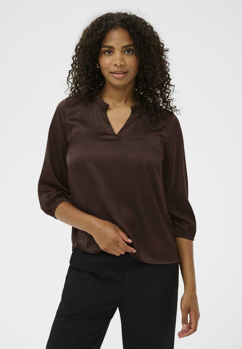Blusa de satén marrón con escote en V y detalle de volantes, mangas 3/4 con puños elásticos, llevada sobre pantalones negros. Textura suave, corte holgado.