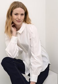 Fehér gombos ing, gallérral, puffos hosszú ujjal és laza fazonnal; sötétkék farmernadrággal párosítva, amelyen látható varrások vannak.