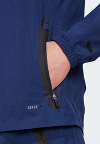 Marineblauwe jas met een gladde textuur, voorzien van een ritssluitingszak, elastische manchetten en "AEROREADY" branding in het zwart.