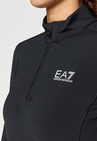 Čierny dlouhý rukáv zipsový top vyrobený z hladkej a elastickej tkaniny, s vysokým golierom a bielym logom EA7 Emporio Armani na hrudi.