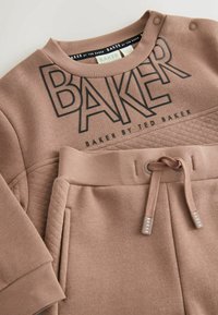 Brązowa bluza i pasujące spodnie. Bluza ma czarny napis "BAKER", pikowaną fakturę oraz napy; spodnie mają elastyczny pasek z sznurkiem.