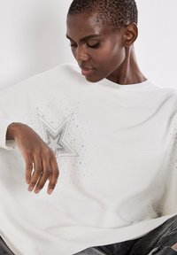 Biały sweter z dzianiny z srebrnym wzorem w gwiazdy ozdobionym kryształkami. Luźny krój i fakturowany materiał z prążkowanym dekoltem.