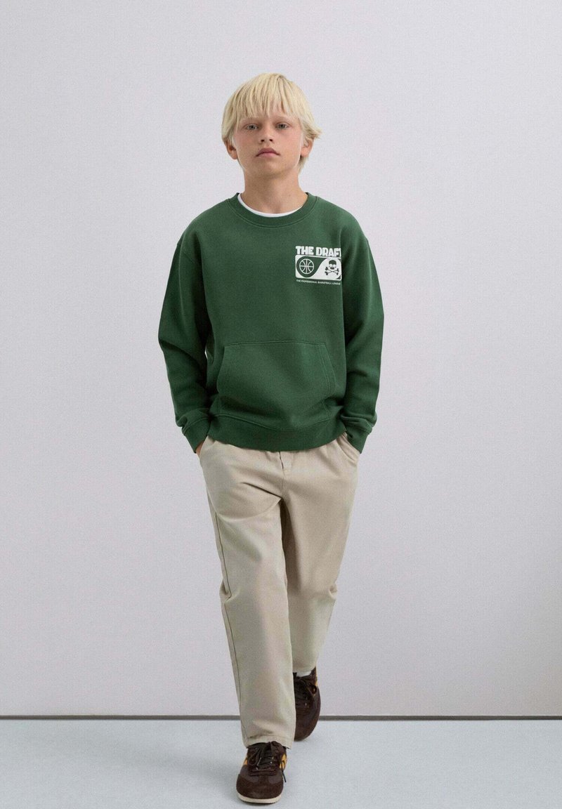 Sudadera verde con bolsillo delantero, detalle de logo en blanco; combinada con pantalones beige claro. El modelo lleva zapatos marrón oscuro. Fondo neutro.