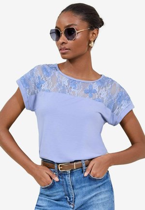 Lichtblauwe T-shirt met een kantoverlay op de schouders, korte opgerolde mouwen en een relaxed fit, gecombineerd met denim en een bruine riem.