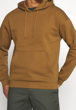 Hombre vistiendo una sudadera con capucha marrón con bolsillo frontal y cordones, combinado con pantalones verde oscuro, de pie con una mano en la cadera.
