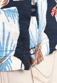 Camisa de manga corta con un patrón floral en azul, marrón y blanco; tejido ligero con costuras visibles y una pequeña etiqueta en el costado.