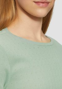 Hellgrünes, geripptes Oberteil mit rundem Halsausschnitt und subtilen textured Mustern mit kleinen Punktdetails. Glatter Stoff mit weicher Verarbeitung.