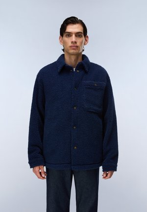 Mann trägt eine dunkelblaue, strukturierte Fleecejacke mit Kragen und Vorderseite Tasche, steht vor einem schlichten hellen Hintergrund.