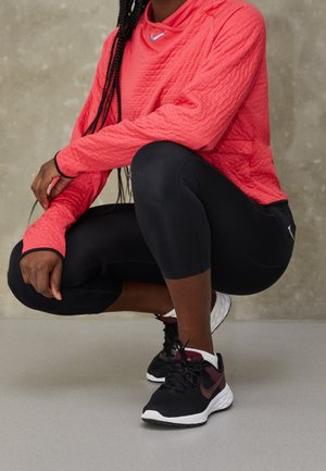 Personne accroupie portant un haut à manches longues texturé corail, des leggings noirs coupés et des chaussures de course Nike noires et bordeaux avec des semelles blanches.